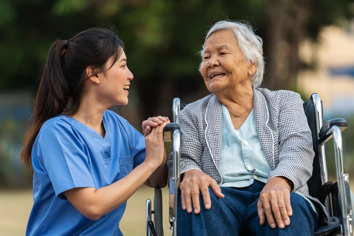 Caregiver holding hands and talking with an older woman in a wheelchair at a rest home in New Zealand.